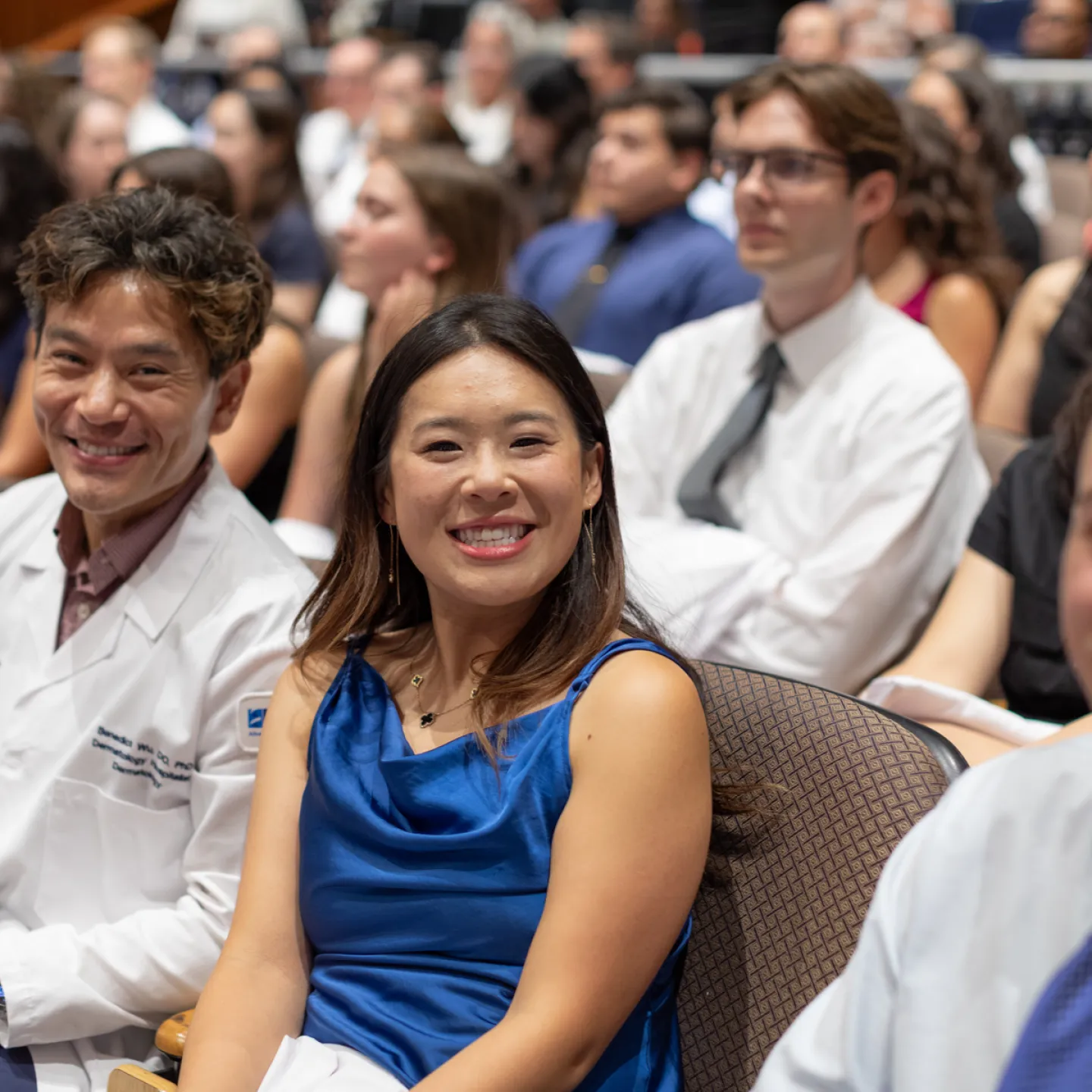 Two audience members, one in a white medical coat, the other in a blue satin dress, smile in their seats.