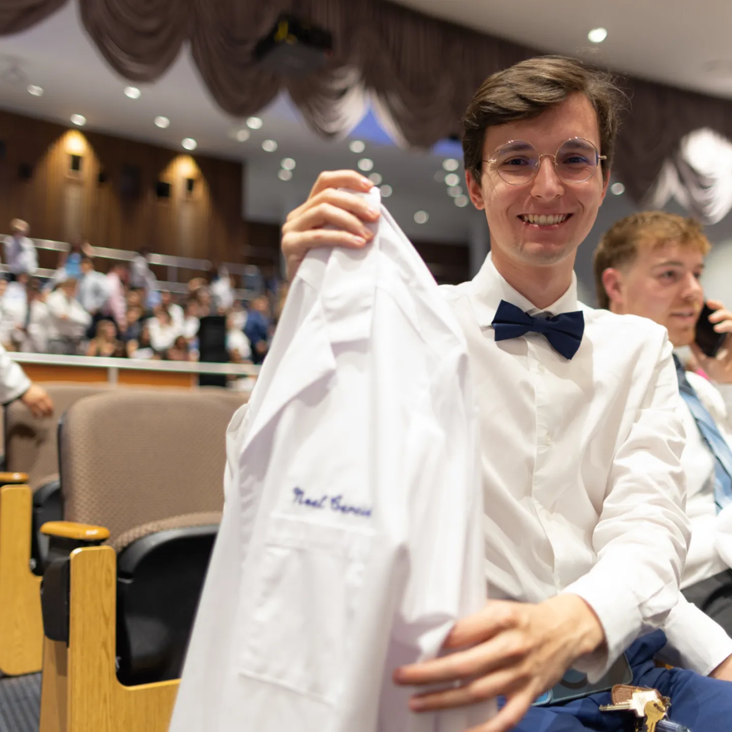A young man in wire-rimmed glasses, a white button down shirt, and a black bowtie lifts his white coat up and smiles. 