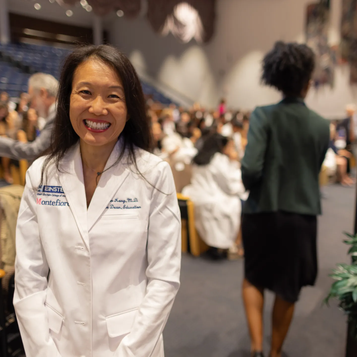 Dr. Yoon Kang, dressed in a white coat, stands and smiles next to the podium at the front of Robbins auditorium.