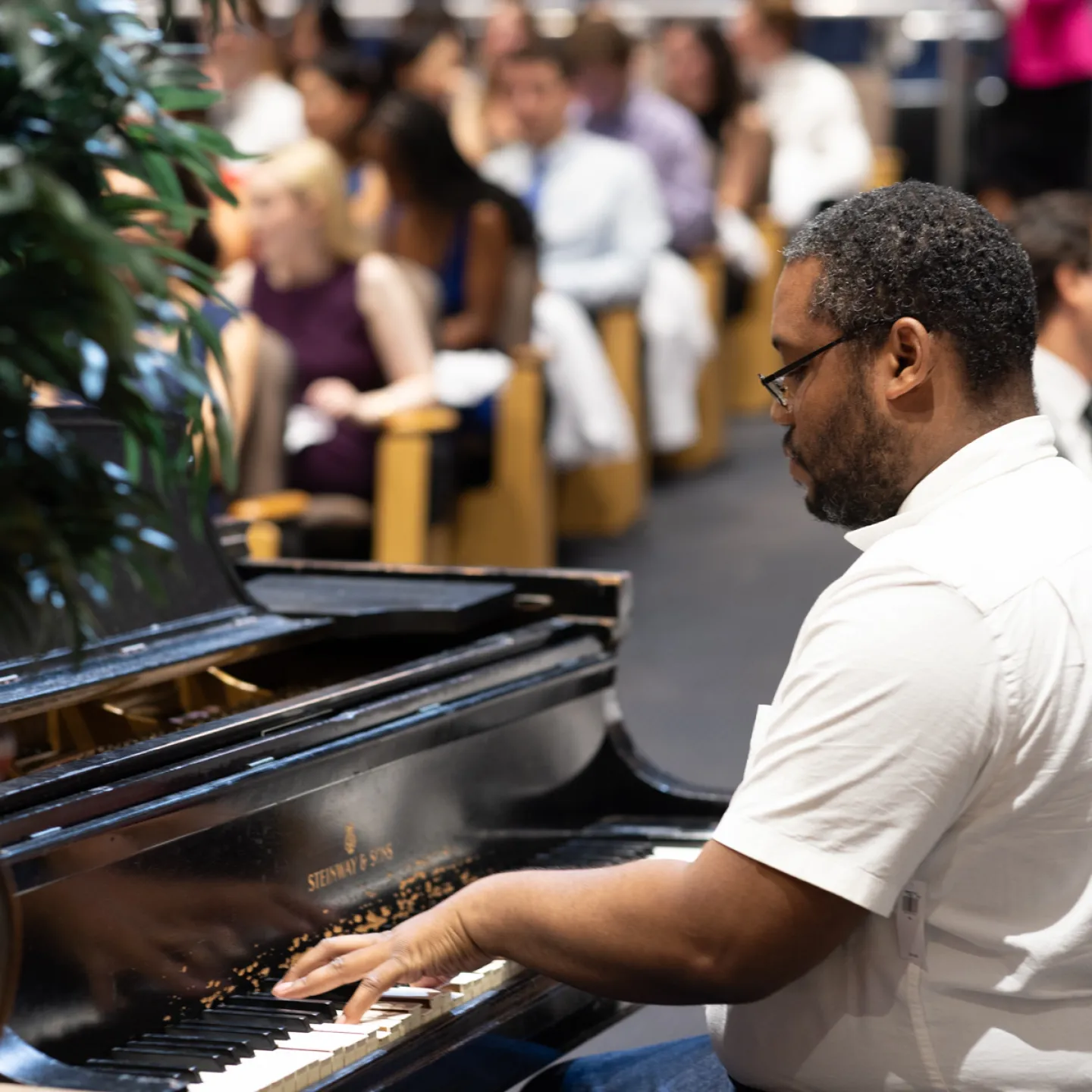 A musician in a white short sleeved button-down shirt plays a black Steinway piano in the front of Robbins auditorium with the audience behind him. 