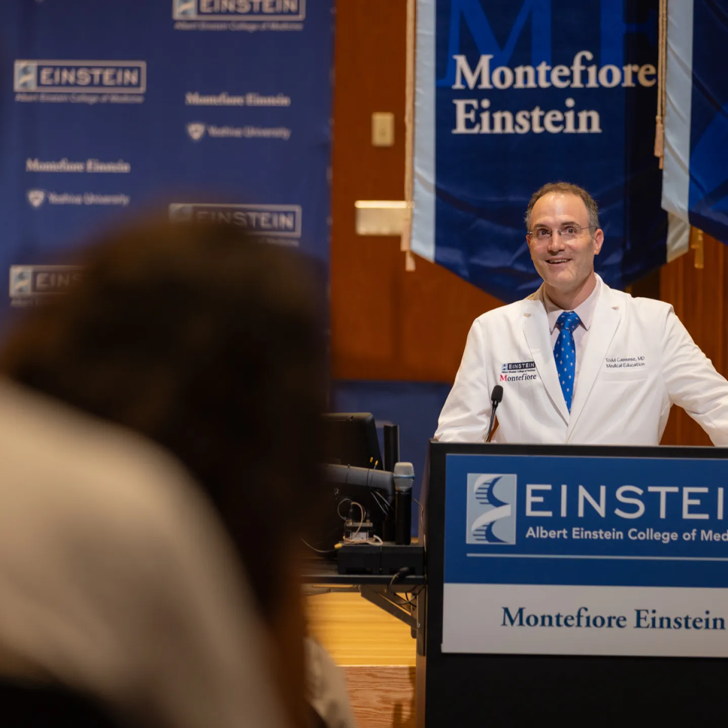 Dr. Todd Cassese, smiling and wearing a white coat and glasses, addresses the audience from the podium in Robbins auditorium, flanked by blue Montefiore Einstein banners. 