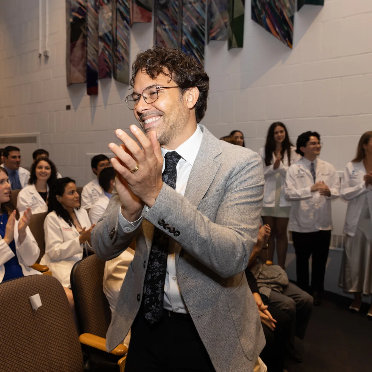 Dr. Correa, dressed in a grey suit jacket, white shirt, black tie, and glasses, stands, smiles, and claps, in Robbins auditorium with students behind him. 