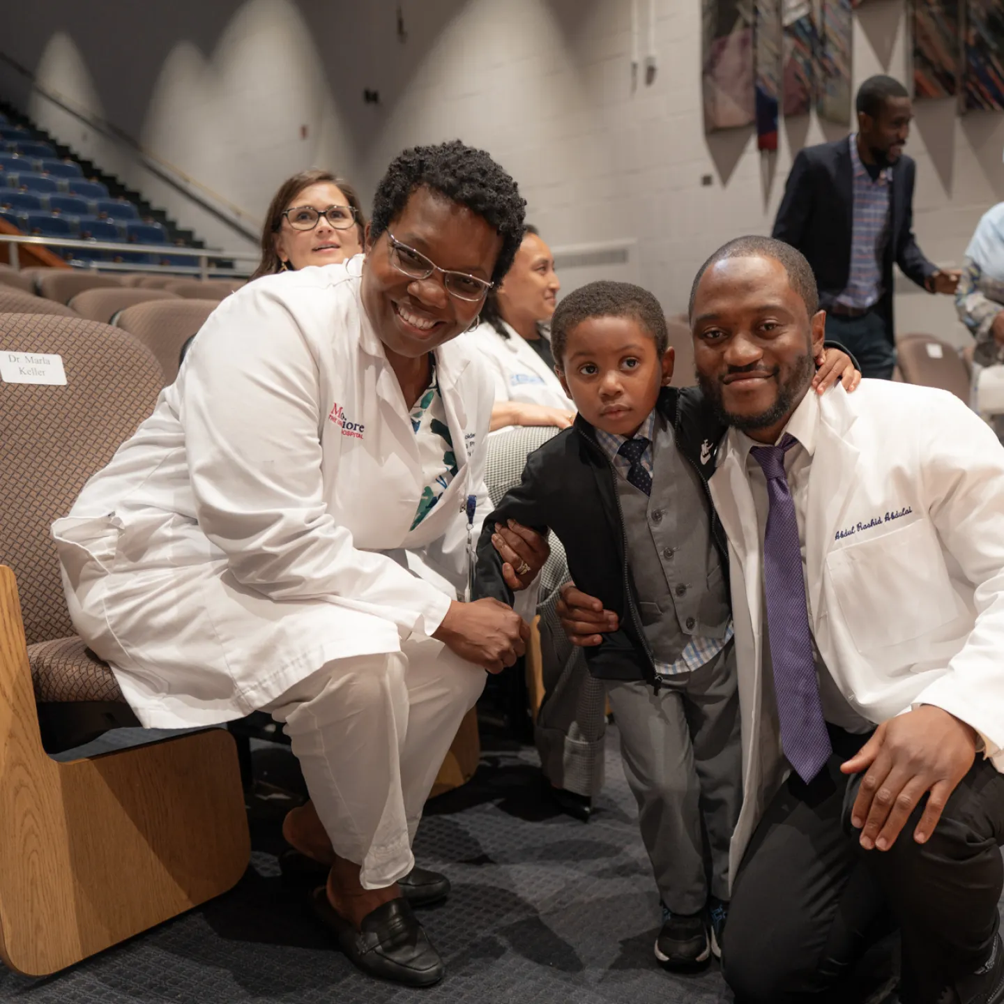 Dr. Holden leans in and smiles with first-year student Abdul Rashid Abdulai. Both are wearing white coats. A young child dressed in jacket, vest, and tie stands between them.