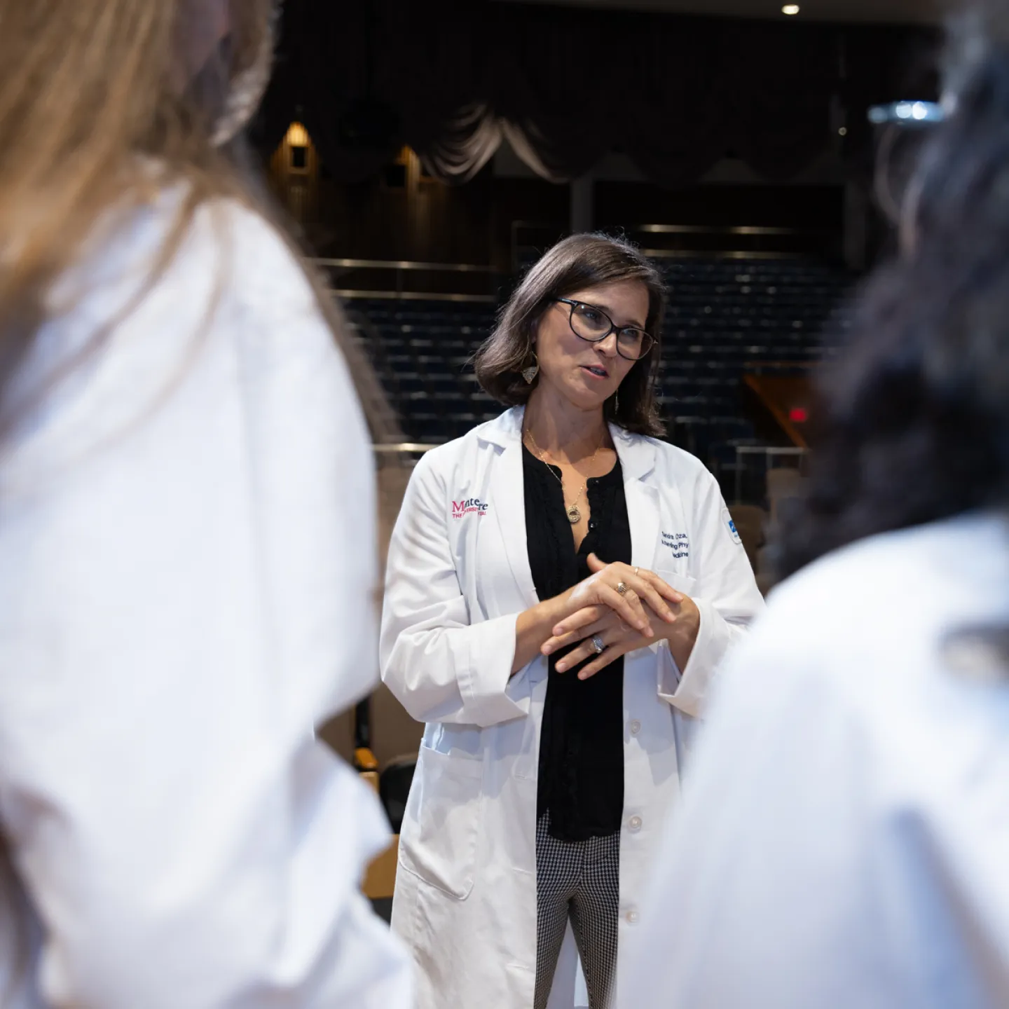 Dr. Oza stands on stage in Robbins auditorium clasping her hands mid-conversation between two medical students with their backs to the camera.