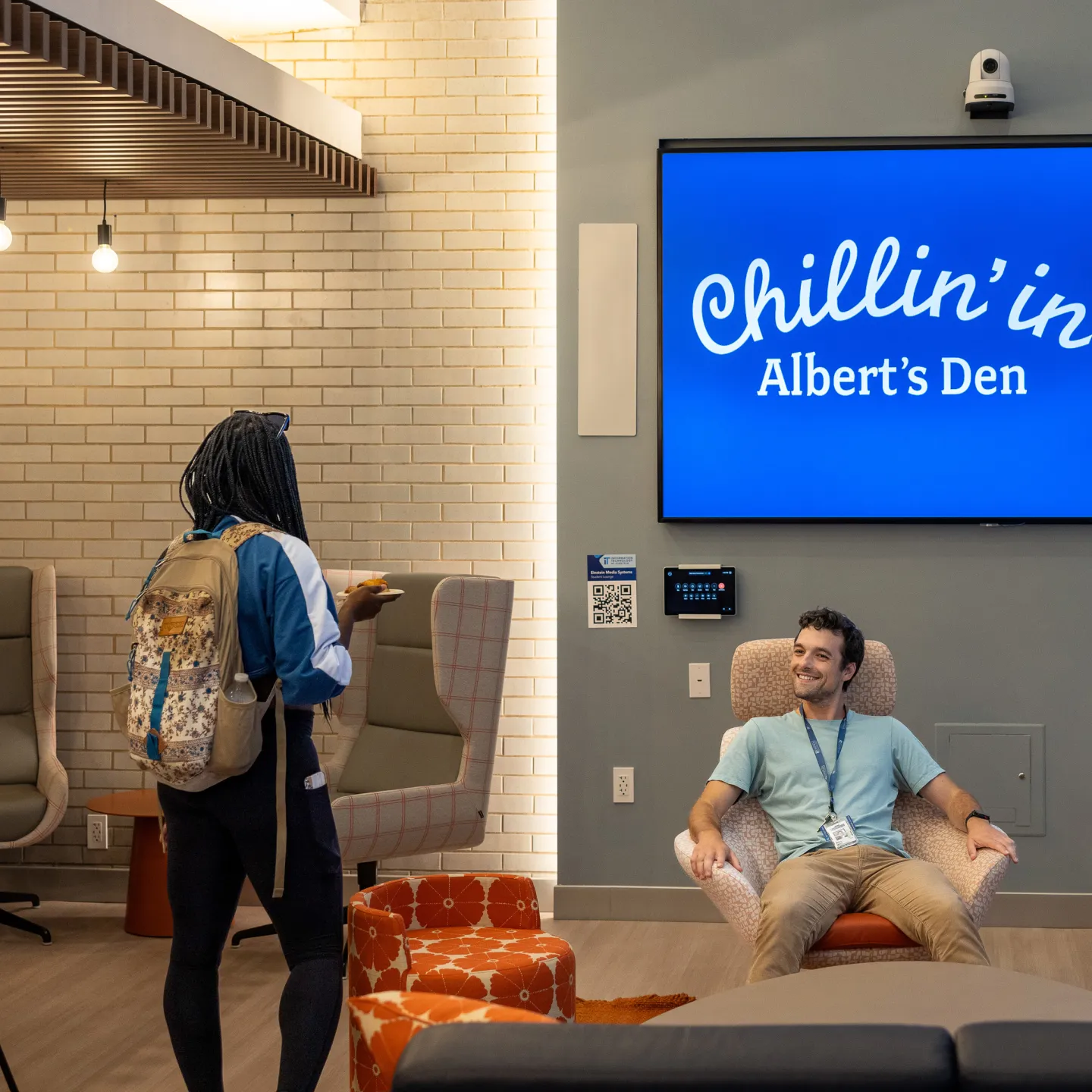 A standing student greets a seated student, smiling and relaxing in a lounge chair below a screen with the message, “Chillin’ in Albert’s Den.”