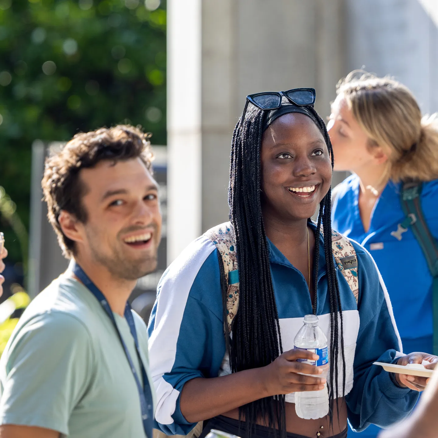 Two students in casual wear smile in a sunny courtyard. They are surrounded by other students relaxing and talking. 