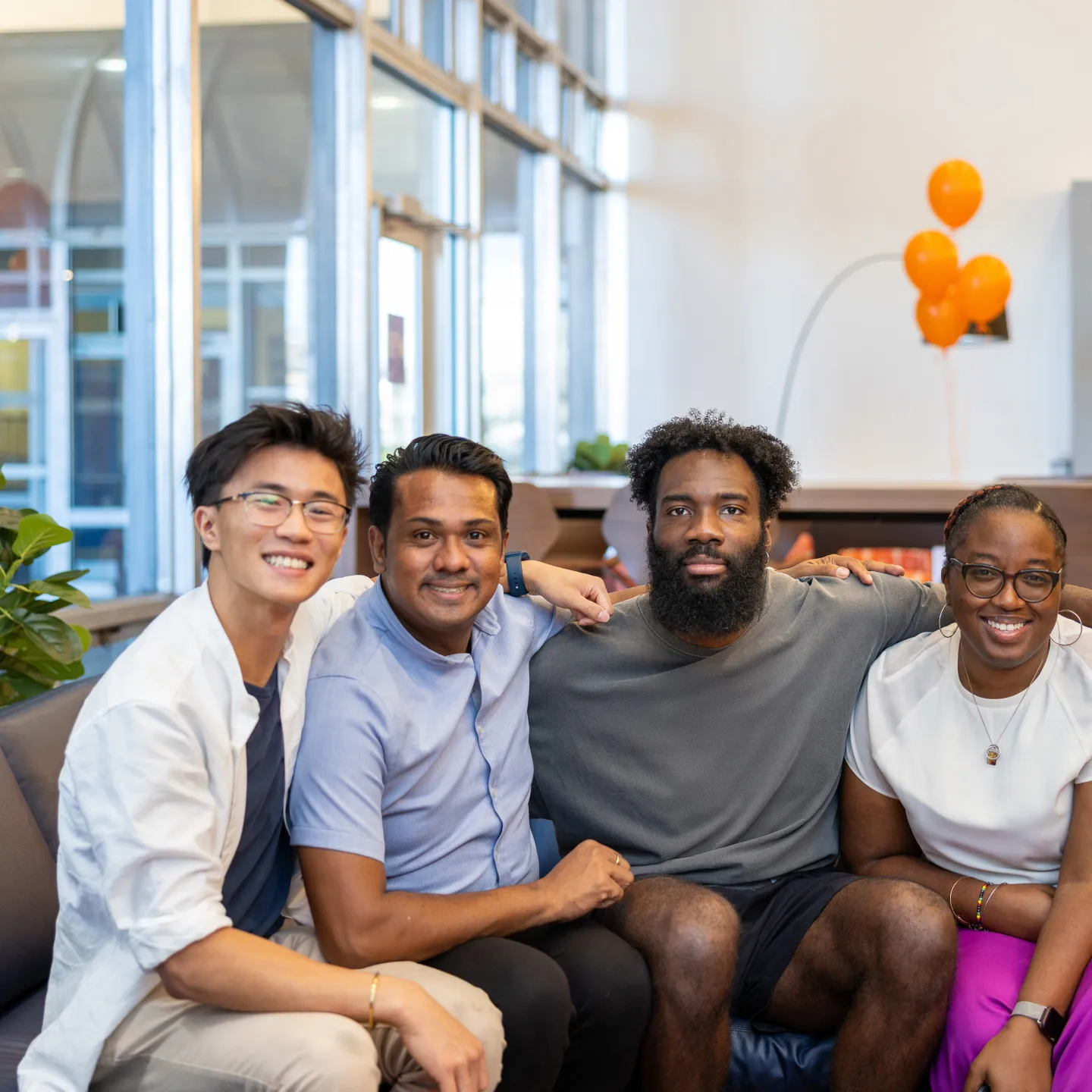 Smiling students and office of student life staff sit on a couch in Albert’s Den surrounded by plants and natural light.