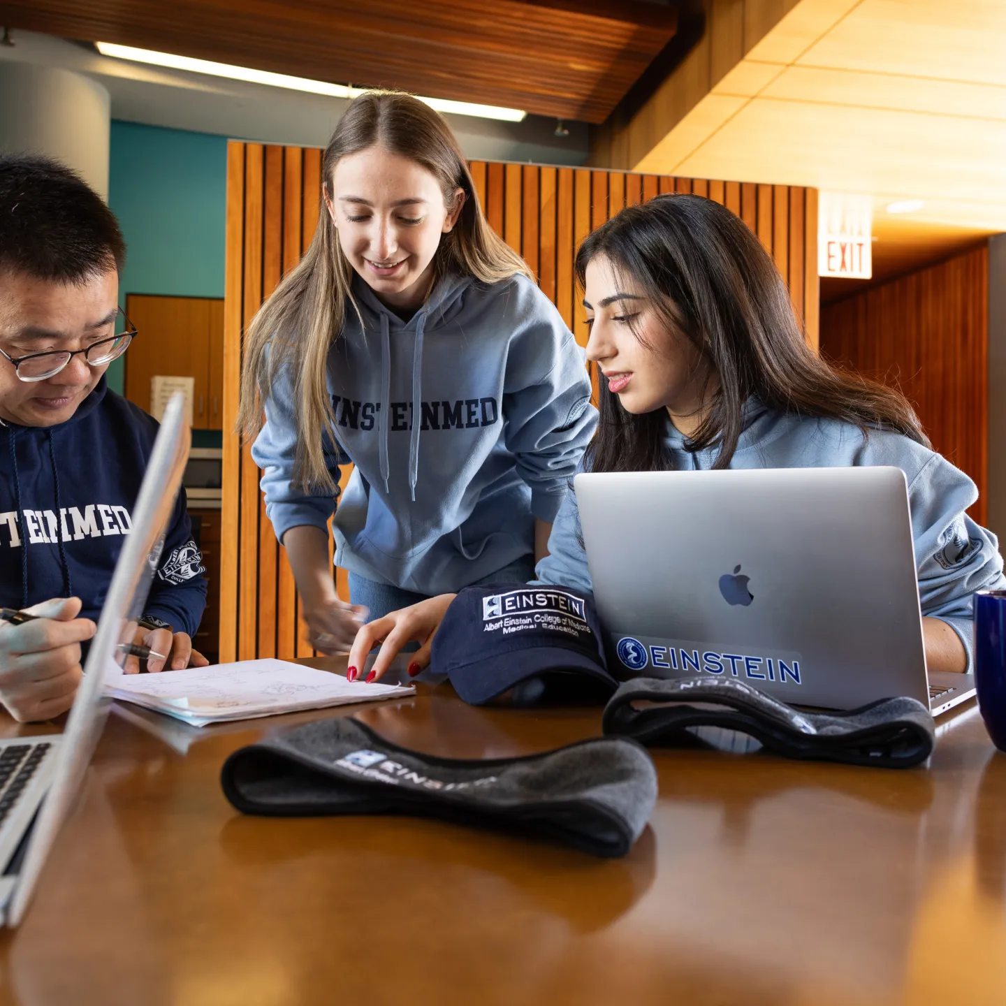 Three students with laptops and notebooks dressed in blue and grey 		Einstein Med sweatshirts gather around a table in Albert’s Den.