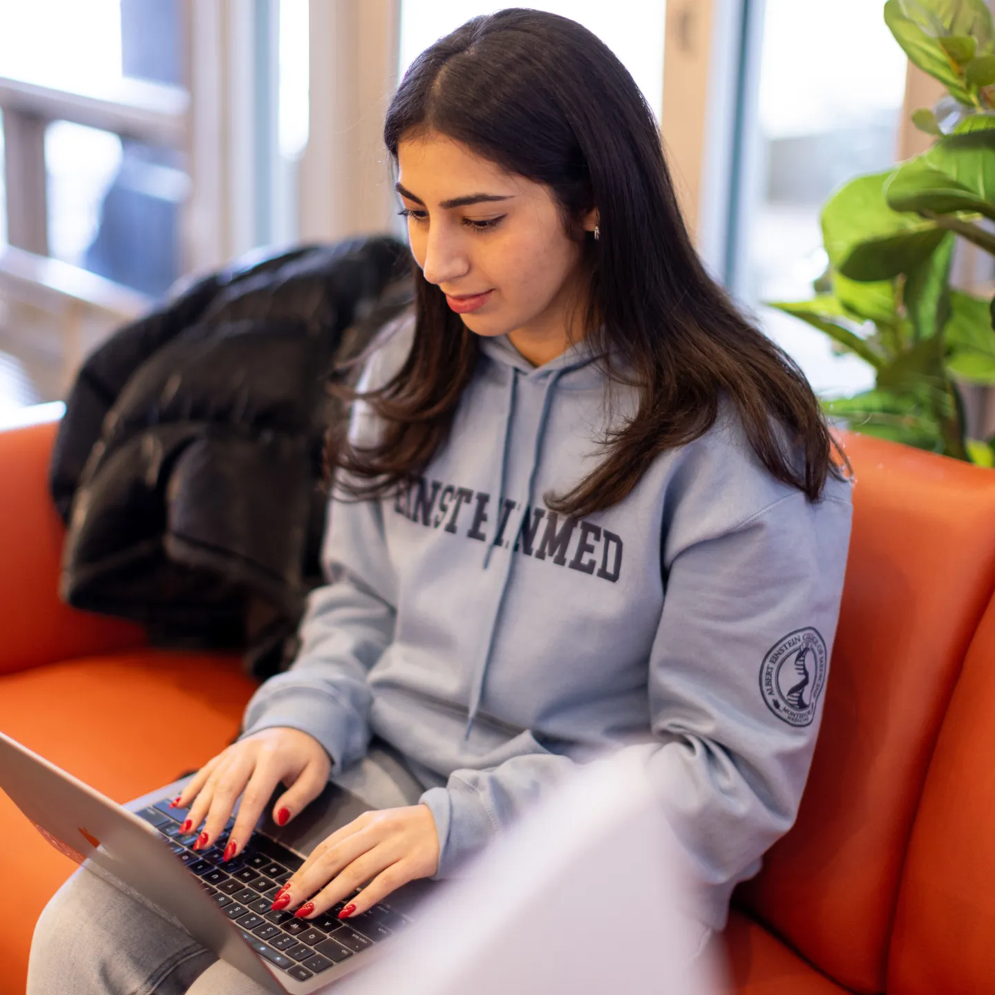 A student in a grey Einstein Med sweatshirt seated on an orange couch works on a laptop with plants in the background. 
