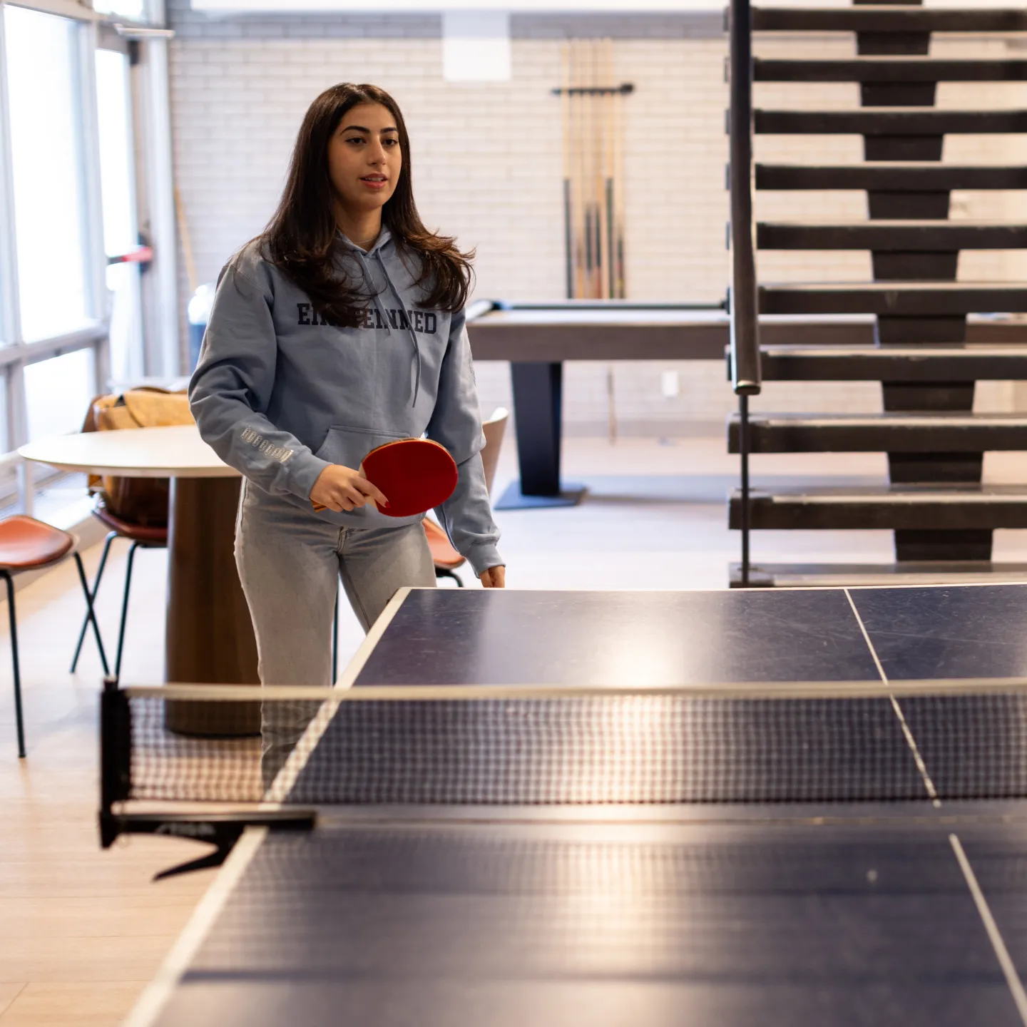 A student in an Einstein Med sweatshirt holds a red paddle and prepares to return a serve at the ping pong table in Albert’s Den. 