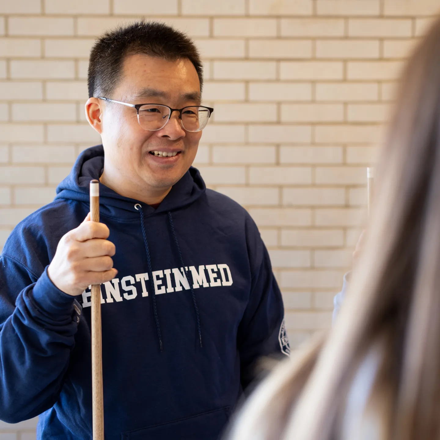 A student in an Einstein Med sweatshirt holds a cue stick and takes a break from a billiards game to talk to another student. 