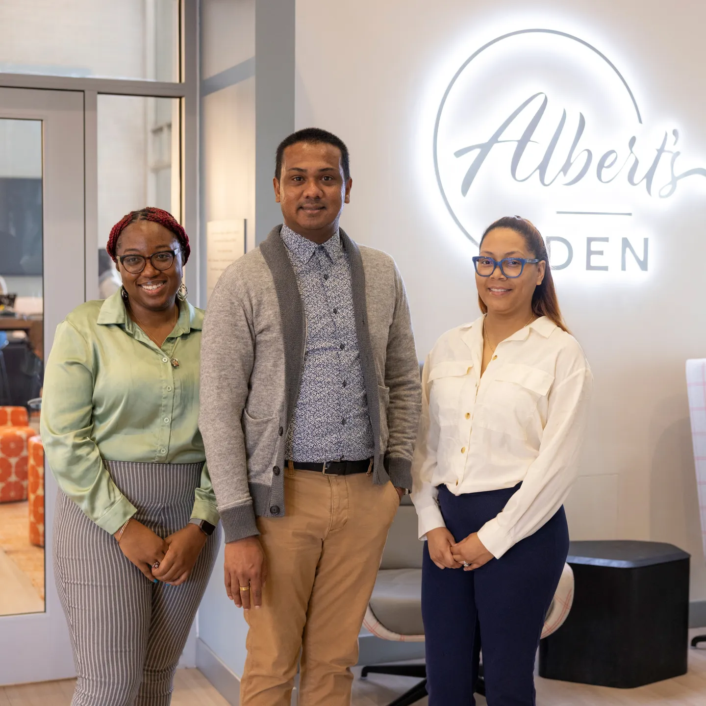 Three staff from Einstein’s office of student life stand smiling with hands crossed in front of a neon sign that says Albert’s Den.