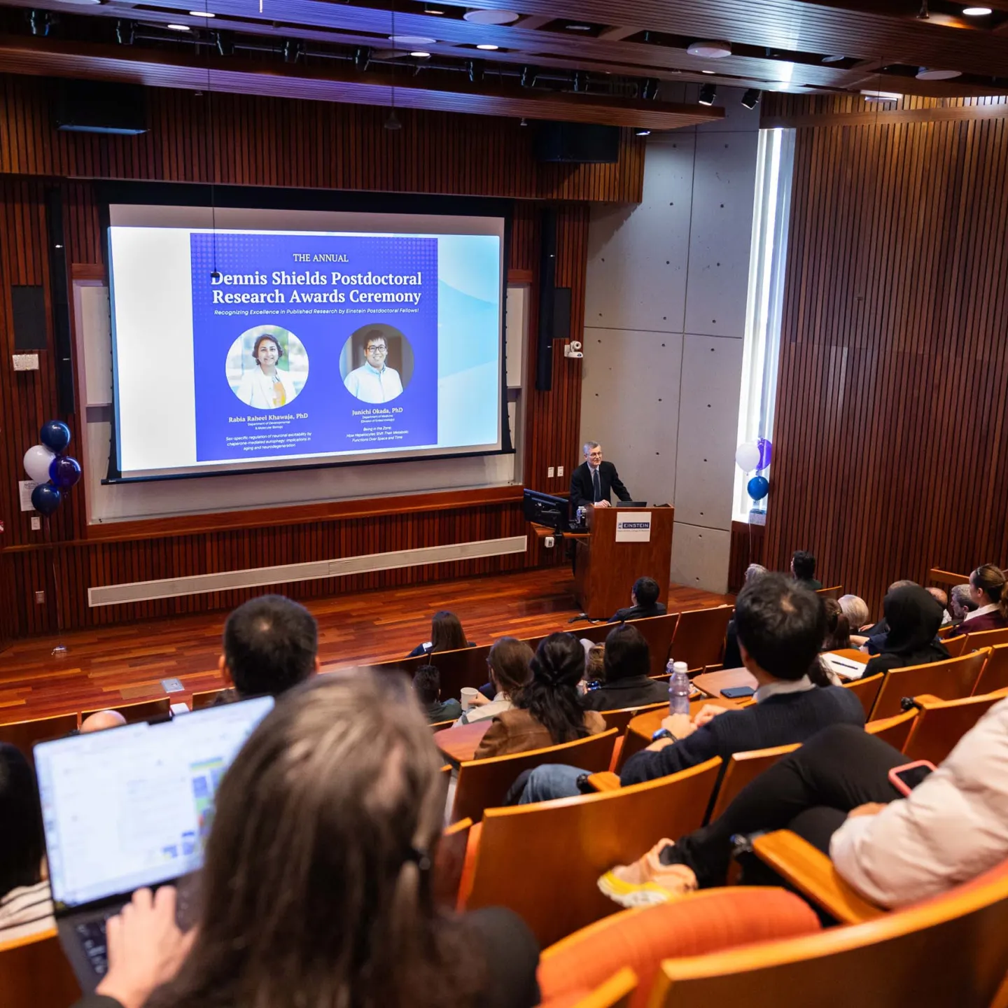 Dean Yaron Tomer at podium speaking in Lefrak Auditorium, panned out image