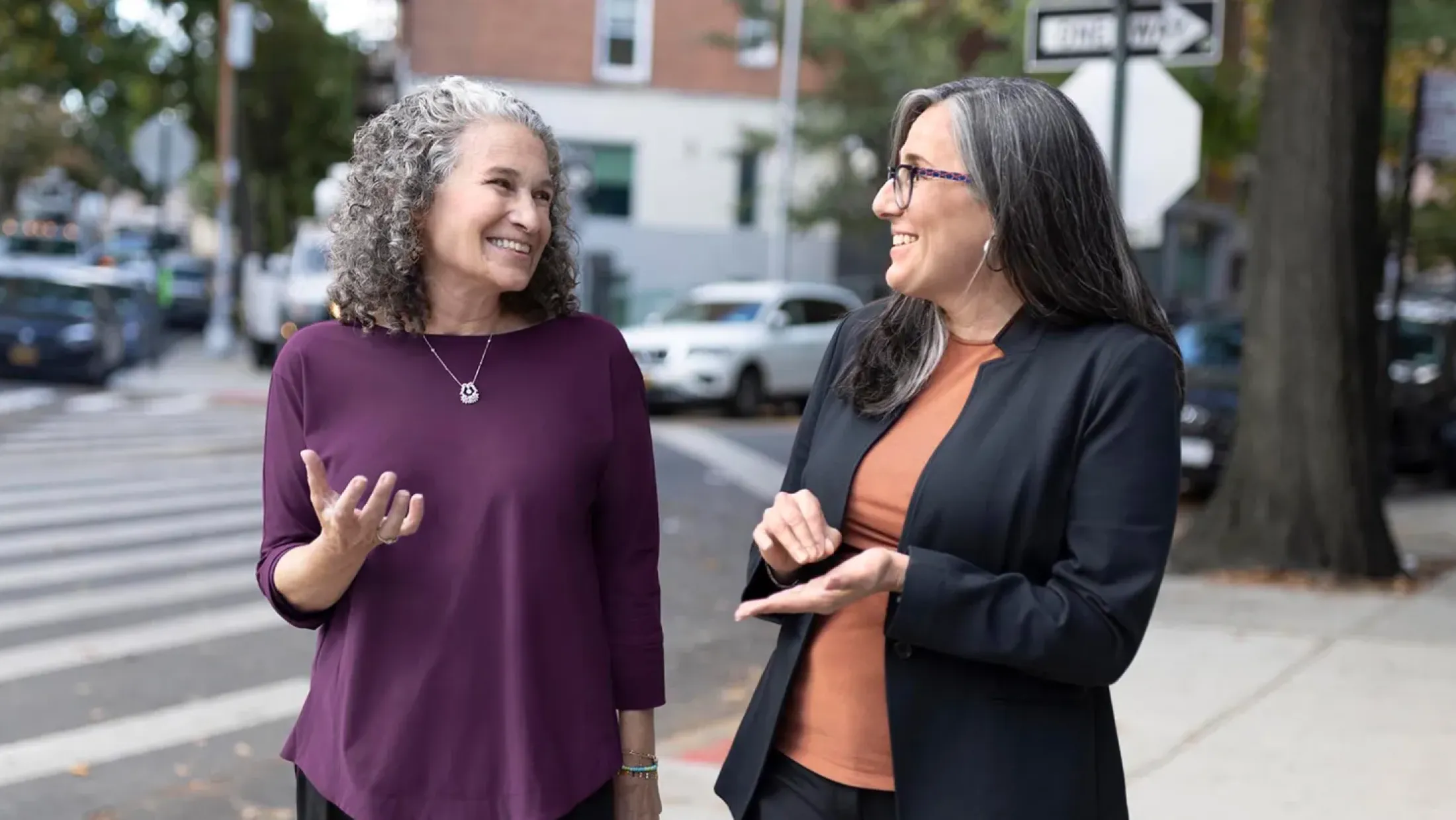 Two women talk outside on a leafy urban street
