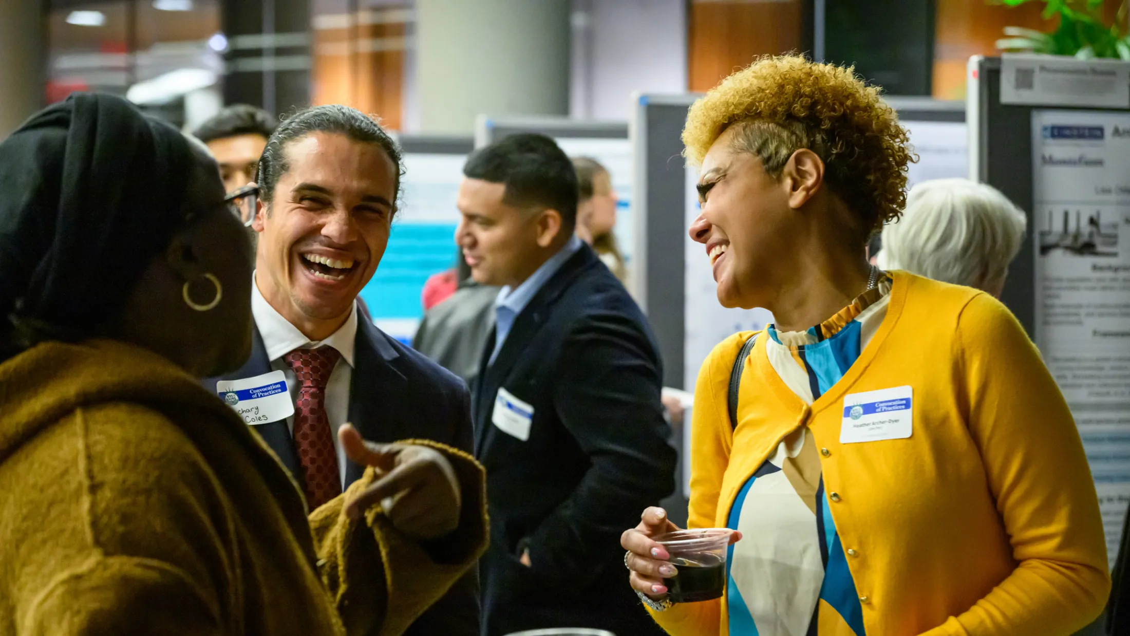 A man in a navy suit and red tie and a woman in a yellow cardigan laugh together beside a row of research posters