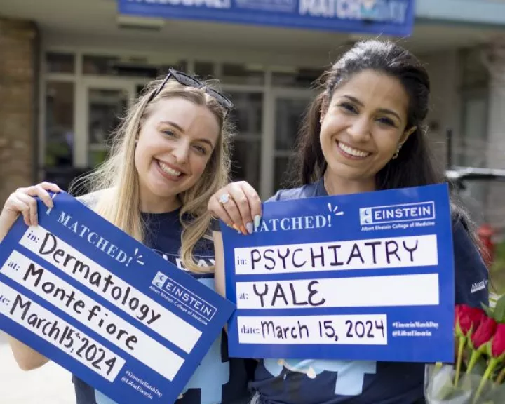 Two fourth-year M.D. students in Match Day t-shirts hold signs announcing that they matched with residency programs in dermatology at Montefiore and psychiatry at Yale