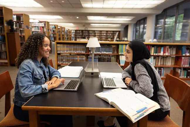 Two students exchange study tips at a desk in the D. Samual Gottesman Library